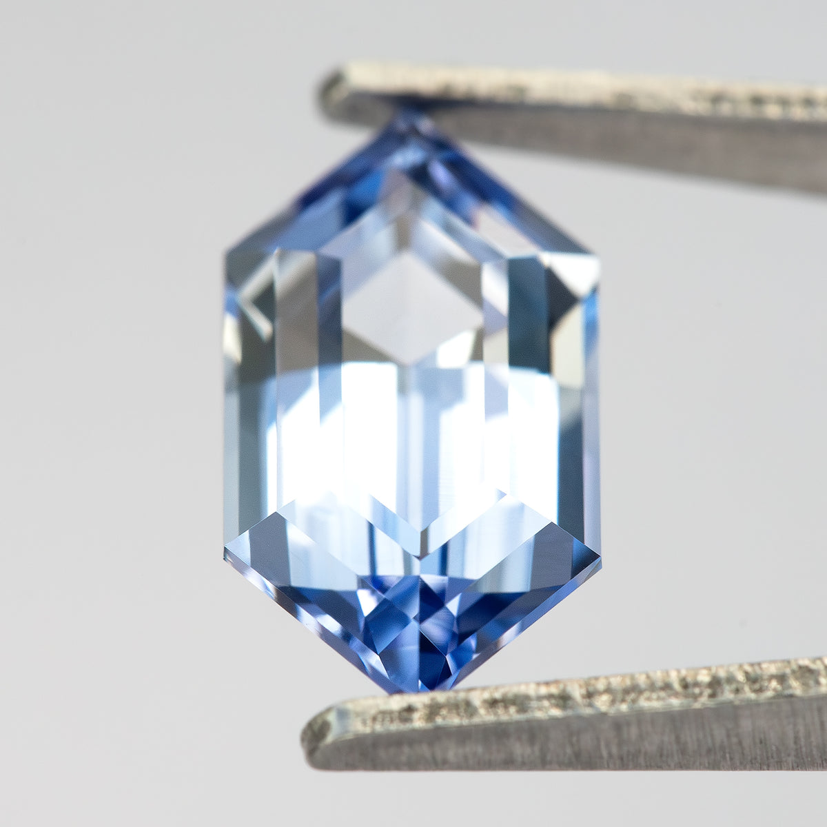 The blue lab-grown sapphire tilted in tweezers, with strong white reflections across the step-cut face.