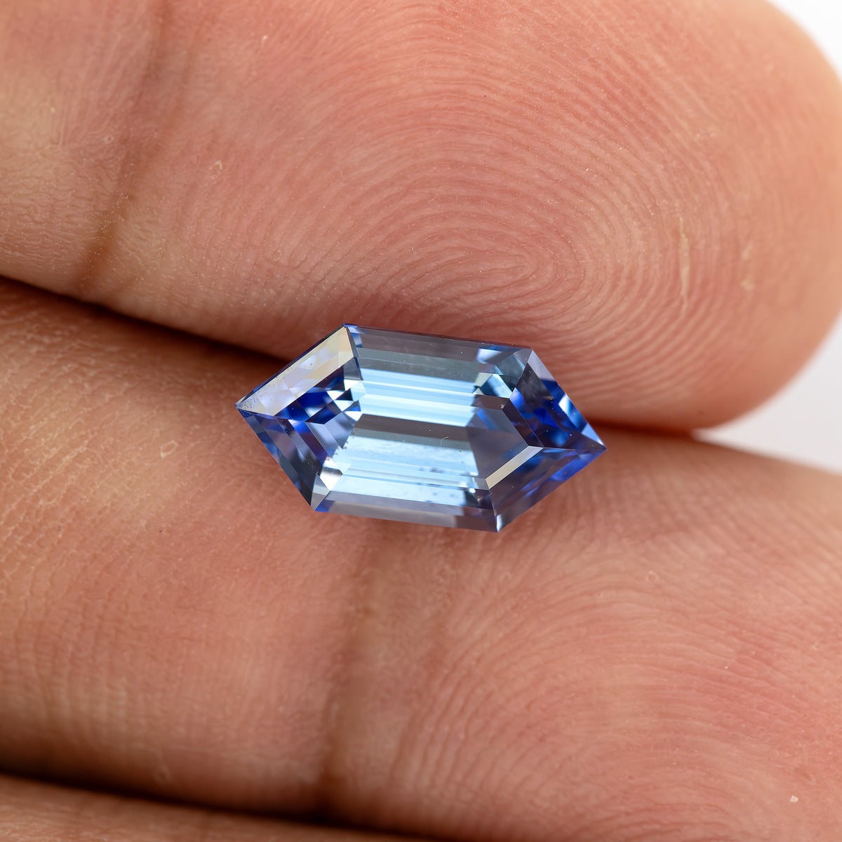 Blue long-hexagon sapphire on fingertips, photographed at a shallow angle to show changing facet reflections.