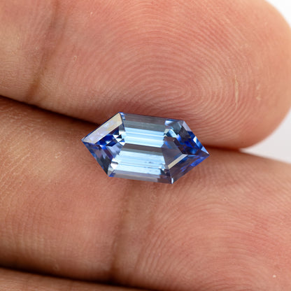 Blue long-hexagon sapphire on fingertips, photographed at a shallow angle to show changing facet reflections.