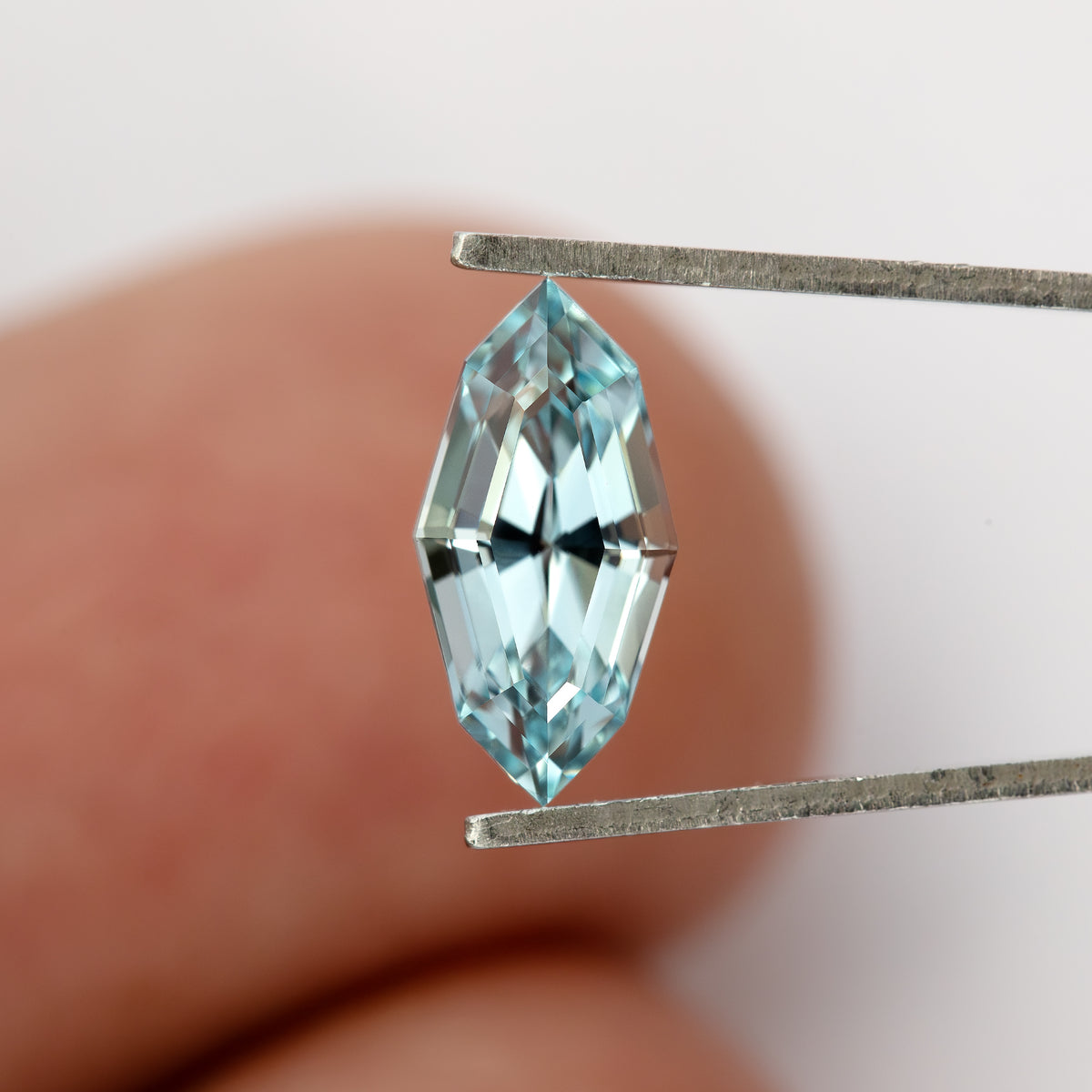 Light blue marquise step-cut lab-grown sapphire face-up vertically in tweezers against a fingertip for scale.