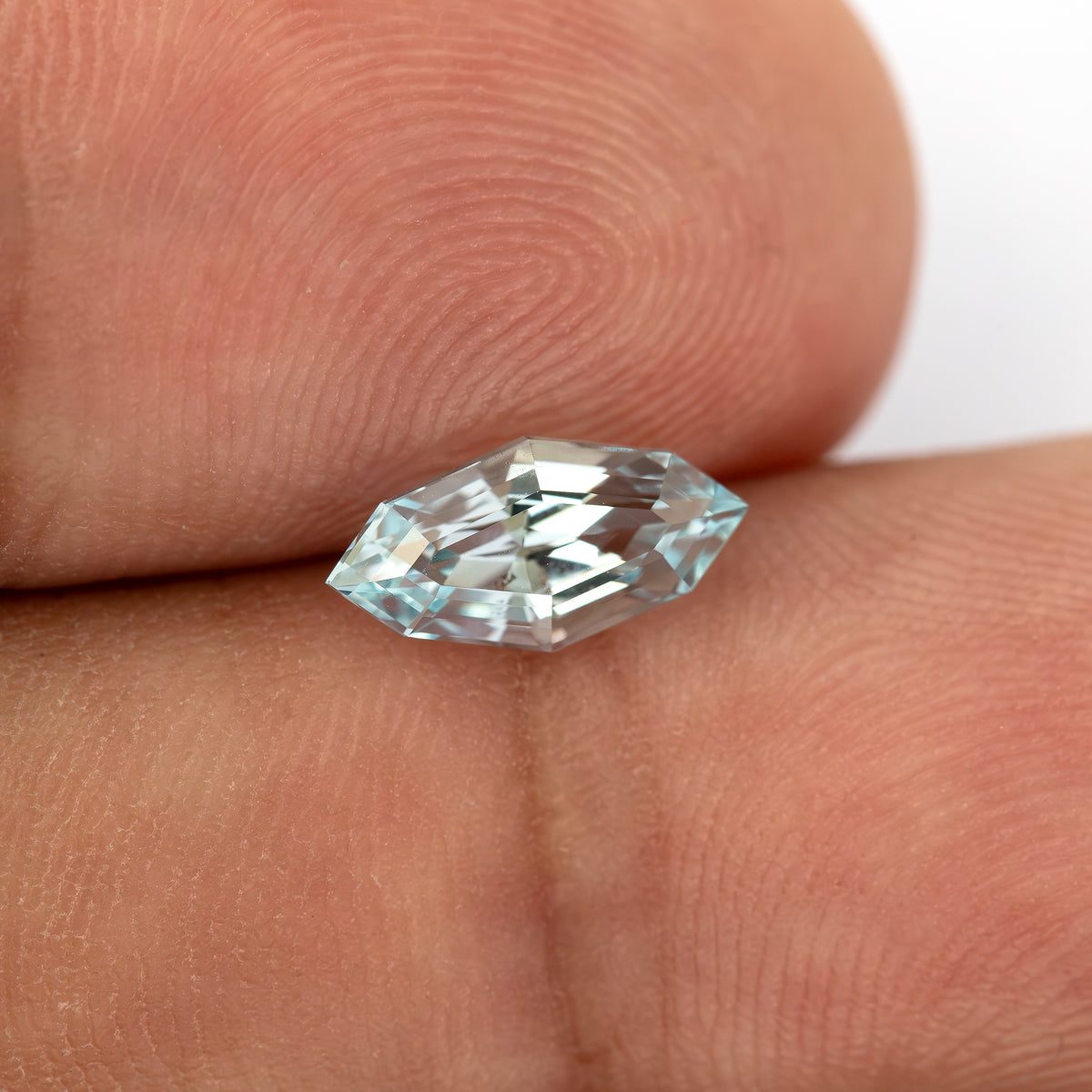 A marquise-shaped, step-cut, light blue lab-grown sapphire resting between human fingers for scale.