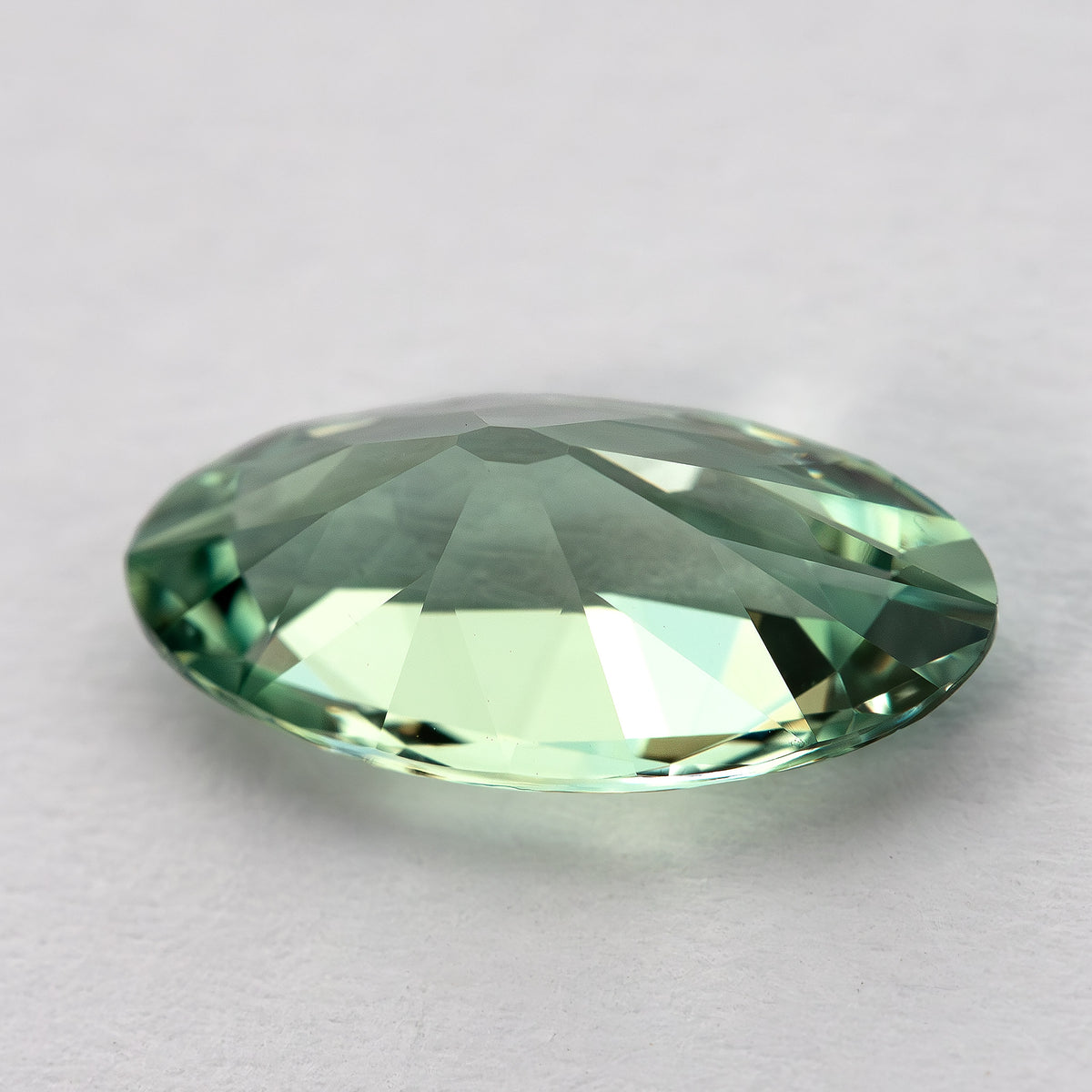 Low angled view of the mint-green lab-grown sapphire resting on a pale surface, showing its thickness and sloping facets.