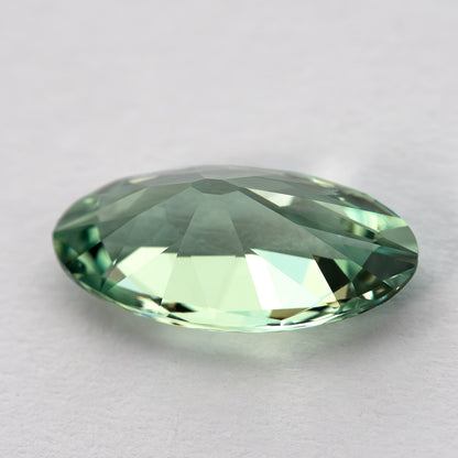 Low angled view of the mint-green lab-grown sapphire resting on a pale surface, showing its thickness and sloping facets.