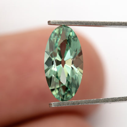 Mint-green lab-grown sapphire face-up in tweezers against a fingertip, giving a sense of scale.