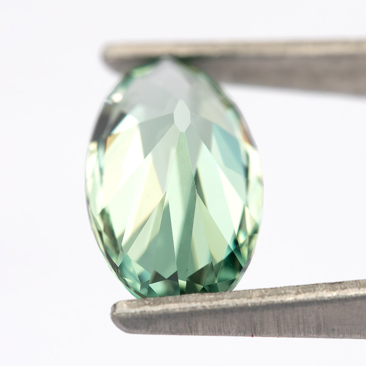 Mint-green lab-grown sapphire tilted backward in tweezers, with a bright pale reflection across the back.