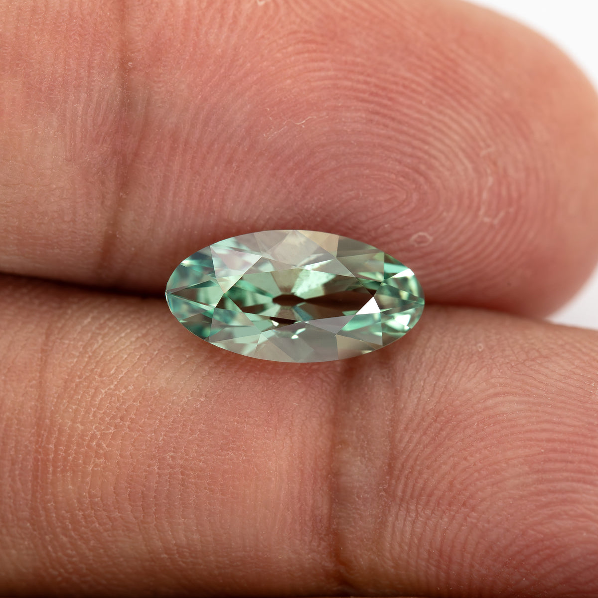 Mint-green lab-grown sapphire lying face-up between fingertips, showing its full outline against skin.