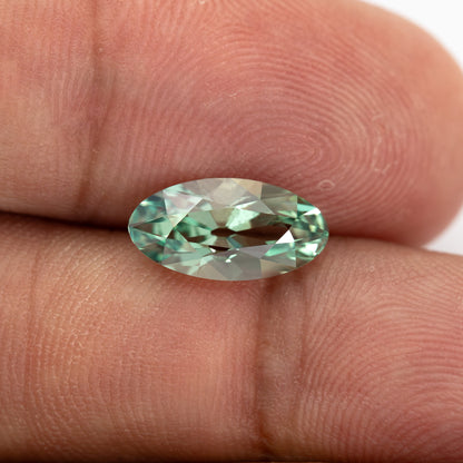Mint-green lab-grown sapphire lying face-up between fingertips, showing its full outline against skin.