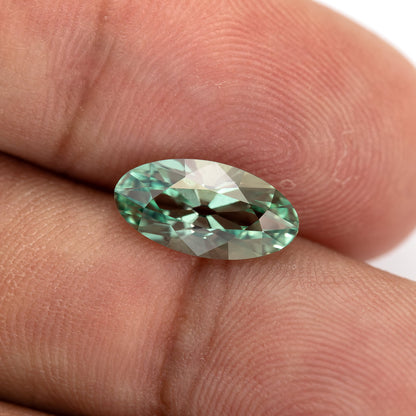 Mint-green lab-grown sapphire held between fingertips and tilted to show contrasting green facet reflections.
