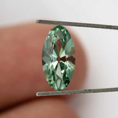 Mint-green lab-grown sapphire upright in tweezers, shown against a fingertip with bright crown reflections.