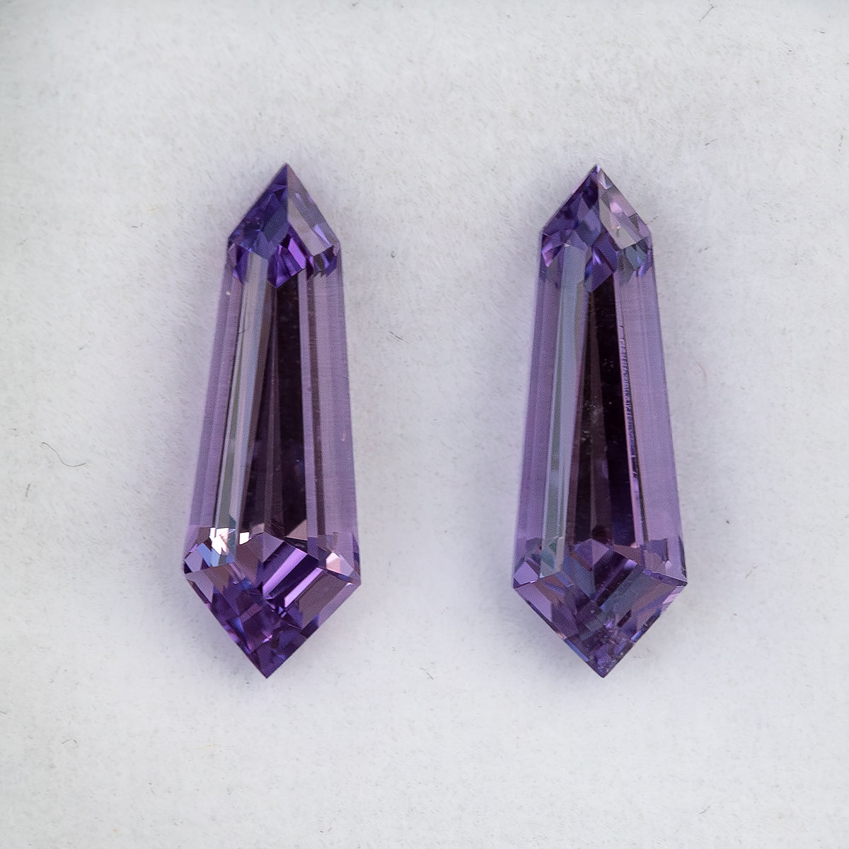 Purple long-shield sapphire pair viewed from the opposite side on a white pad, with long dark central reflections.