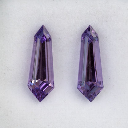 Purple long-shield sapphire pair viewed from the opposite side on a white pad, with long dark central reflections.