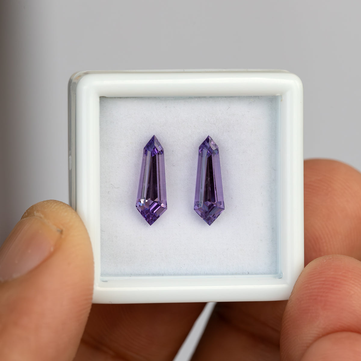 Two purple long-shield lab-grown sapphires in a white gem box, with a thumb visible beside it for scale.