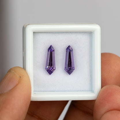 Two purple long-shield lab-grown sapphires in a white gem box, with a thumb visible beside it for scale.