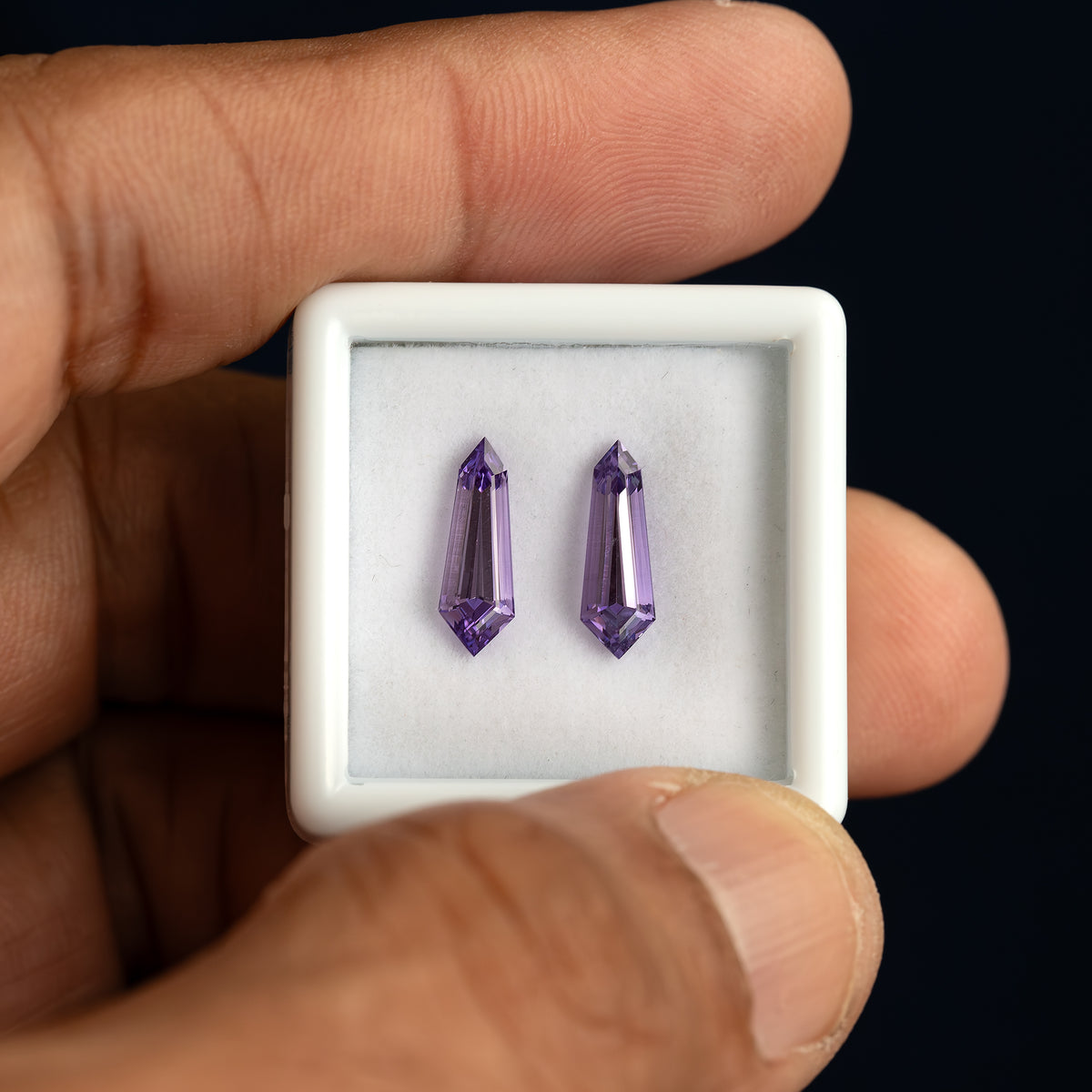 A hand holding a white gem box containing the purple long-shield sapphire pair.