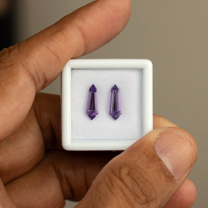 White gem box held between fingers, showing the purple long-shield sapphire pair inside.