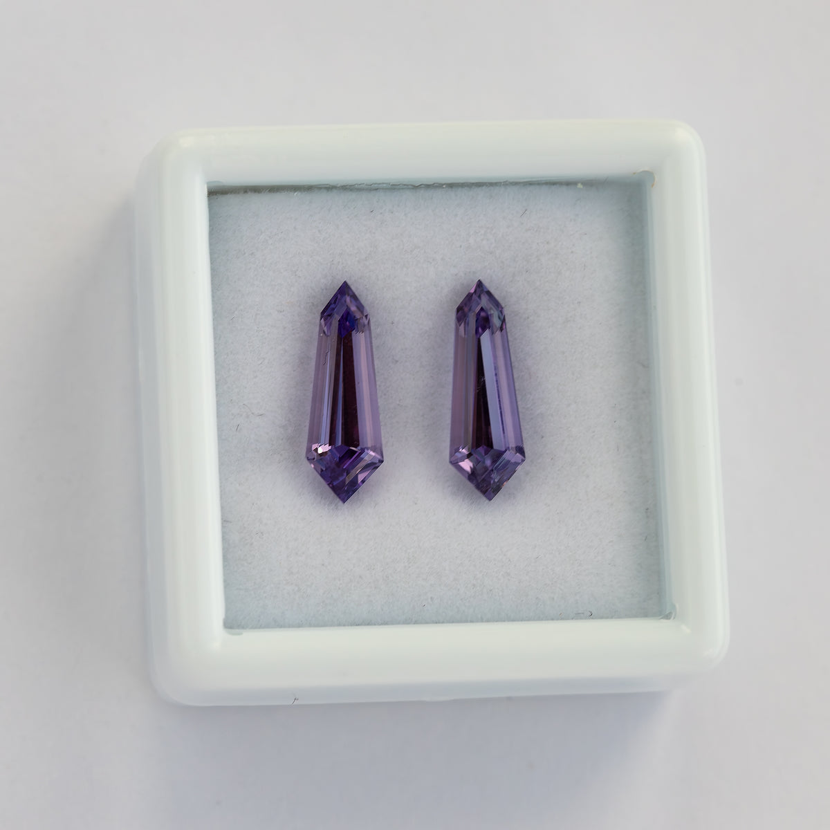 Two purple long-shield sapphires upright inside a white padded gem box on a pale surface.