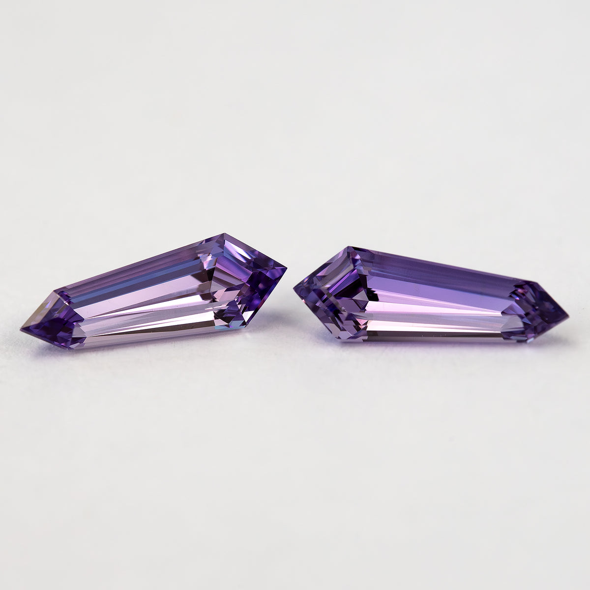 Two purple long-shield lab-grown sapphires at an angle on white, showing their broad tables and step-cut crown facets.