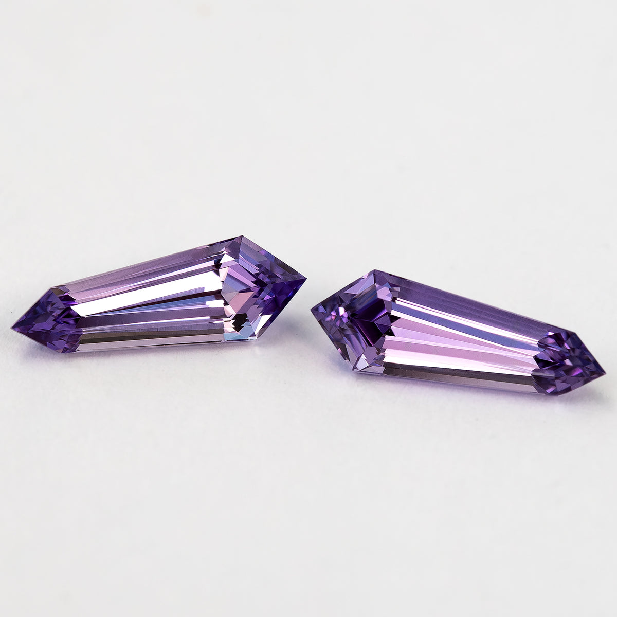 The purple long-shield sapphire pair at a shallow diagonal angle, with pale reflections across both step-cut faces.