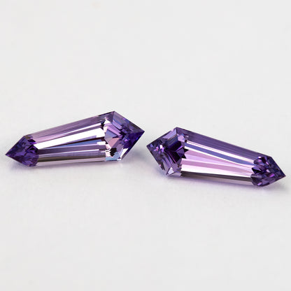 The purple long-shield sapphire pair at a shallow diagonal angle, with pale reflections across both step-cut faces.
