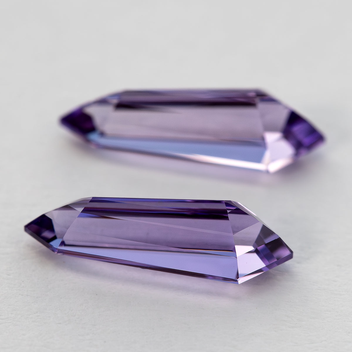 Low parallel side view of the purple long-shield sapphire pair, showing their depth and stepped edges.