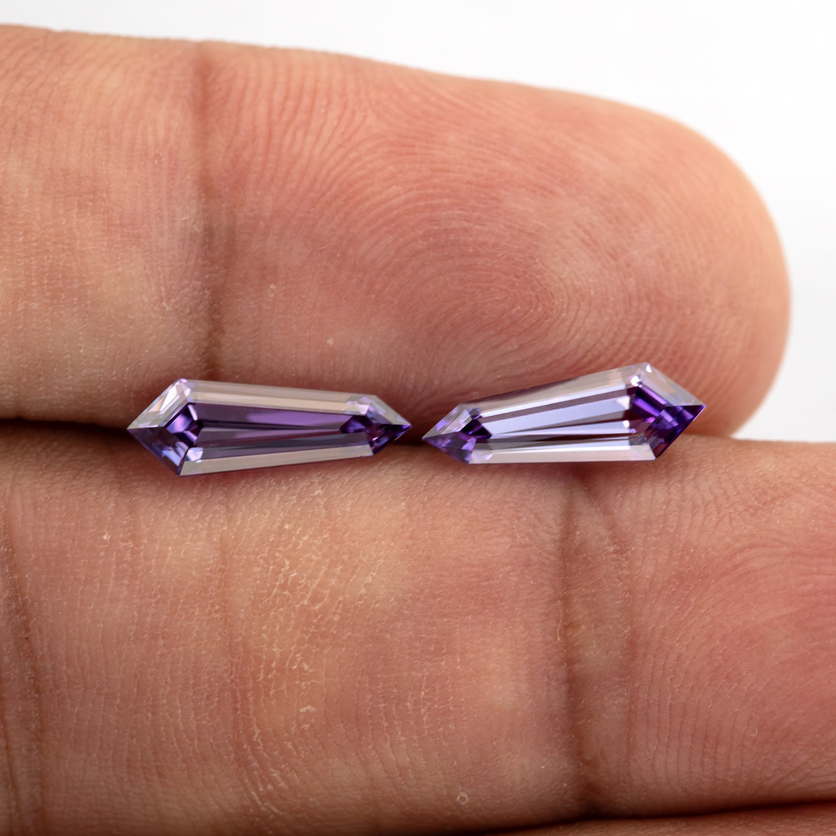 Two purple long-shield lab-grown sapphires resting side by side on fingertips, with their step-cut faces visible.