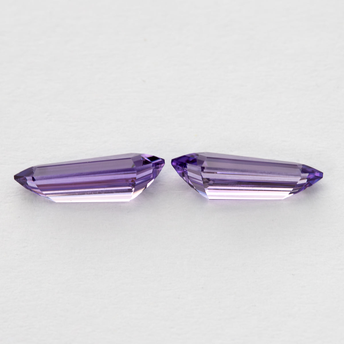 Low opposite-side view of the purple long-shield sapphire pair, with pale reflections along their long facets.