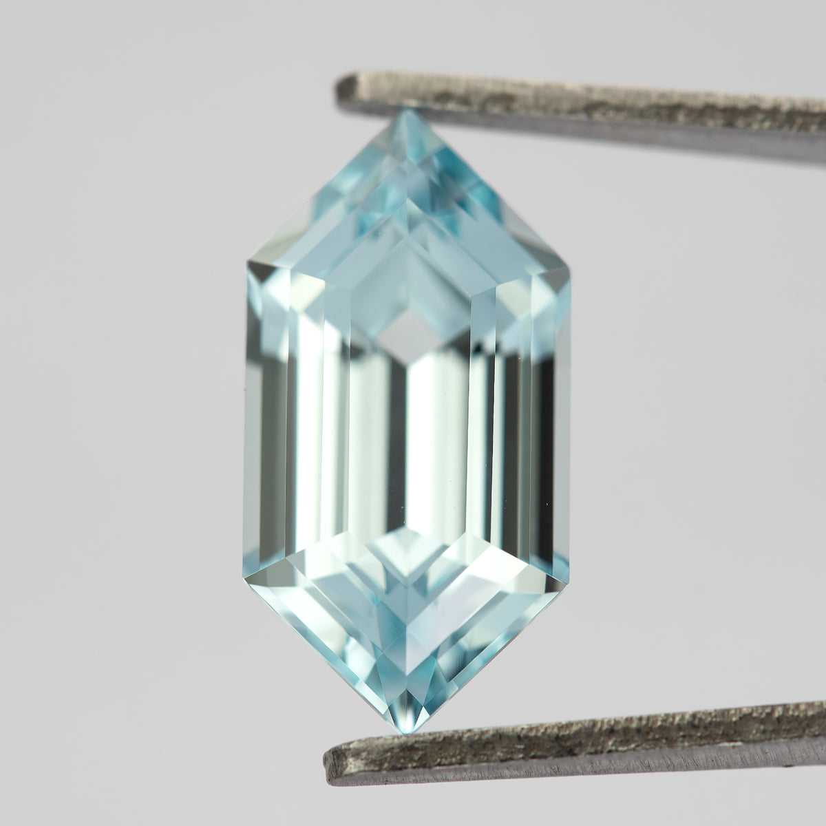 Pale aqua-blue lab-grown sapphire held upright in tweezers, showing its pointed six-sided outline and long step facets.