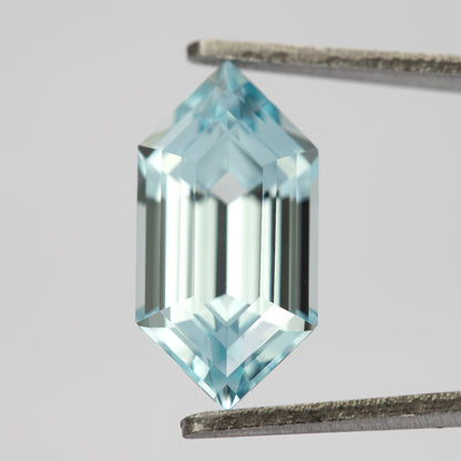Pale aqua-blue lab-grown sapphire held upright in tweezers, showing its pointed six-sided outline and long step facets.