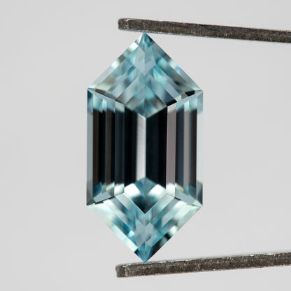 Aqua-blue lab-grown sapphire tilted in tweezers, showing its pointed ends and dark central step reflections.