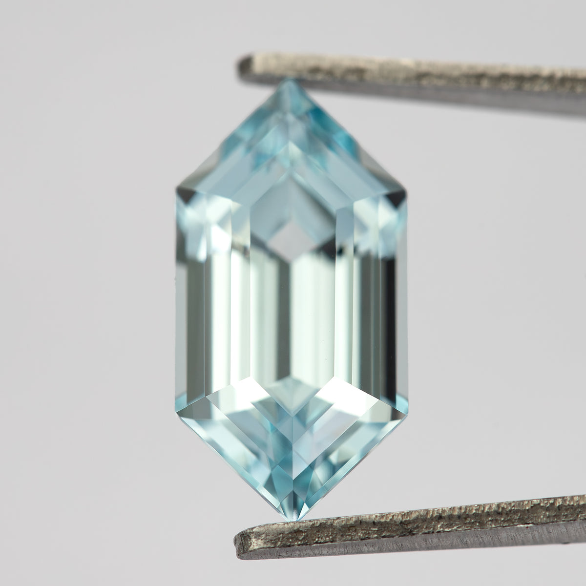 Pale aqua-blue lab-grown sapphire upright in tweezers, with a broad bright reflection across the central steps.