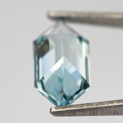 Aqua-blue lab-grown sapphire viewed obliquely from the back in tweezers, revealing the pointed pavilion.