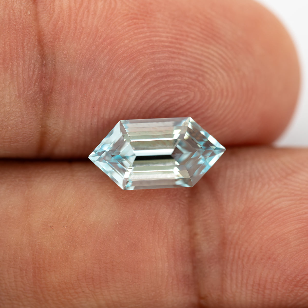 Aqua-blue lab-grown sapphire resting horizontally between fingertips, showing the full six-sided outline for scale.