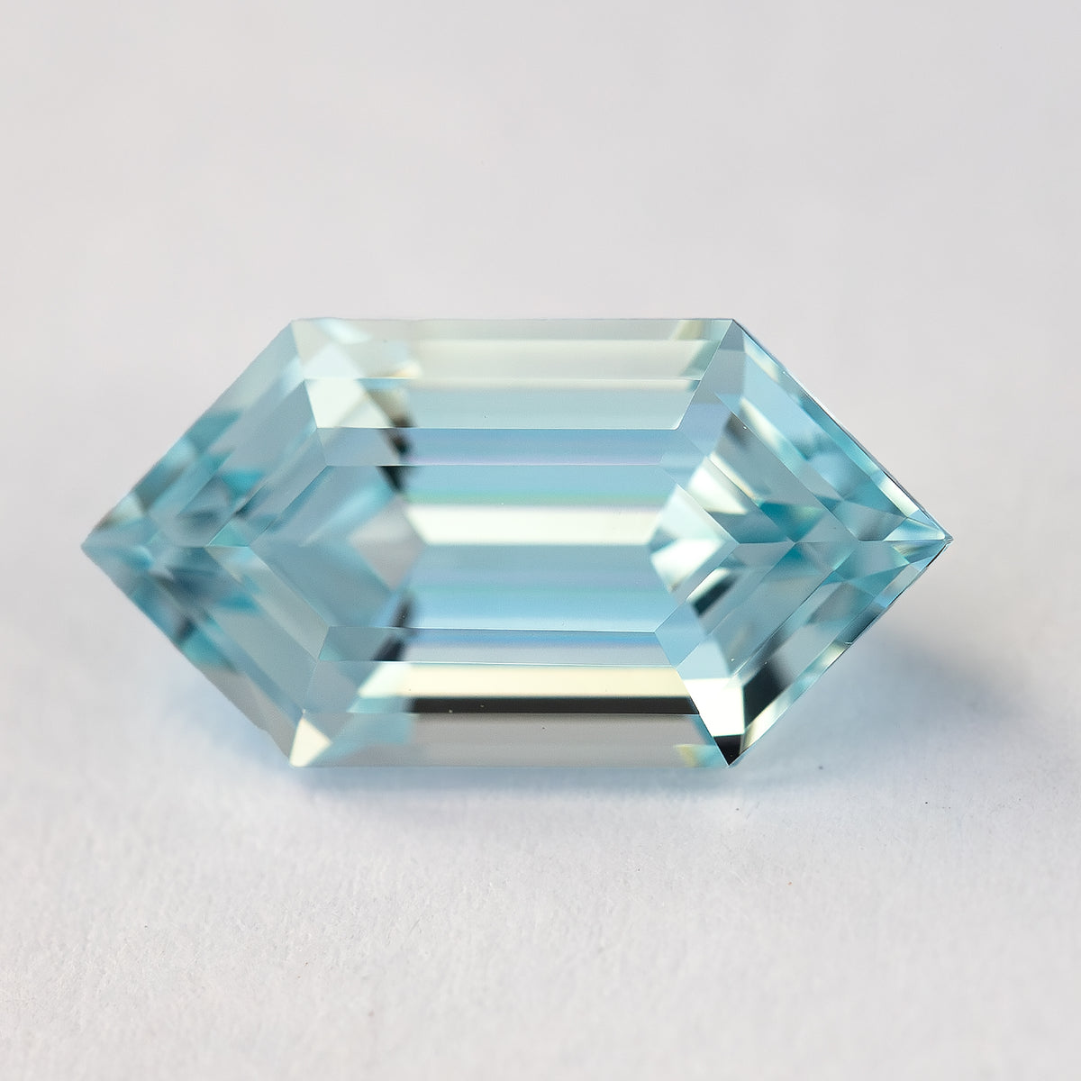Horizontal face-up macro of the aqua-blue lab-grown sapphire on a pale surface, showing its hexagonal outline and long step facets.