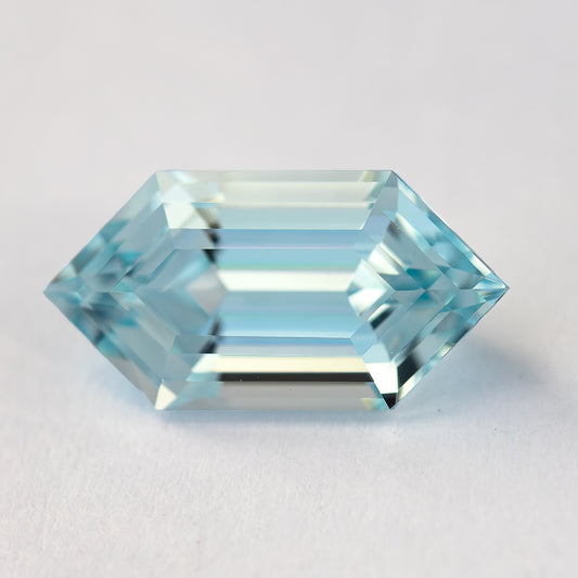 Horizontal face-up macro of the aqua-blue lab-grown sapphire on a pale surface, showing its hexagonal outline and long step facets.