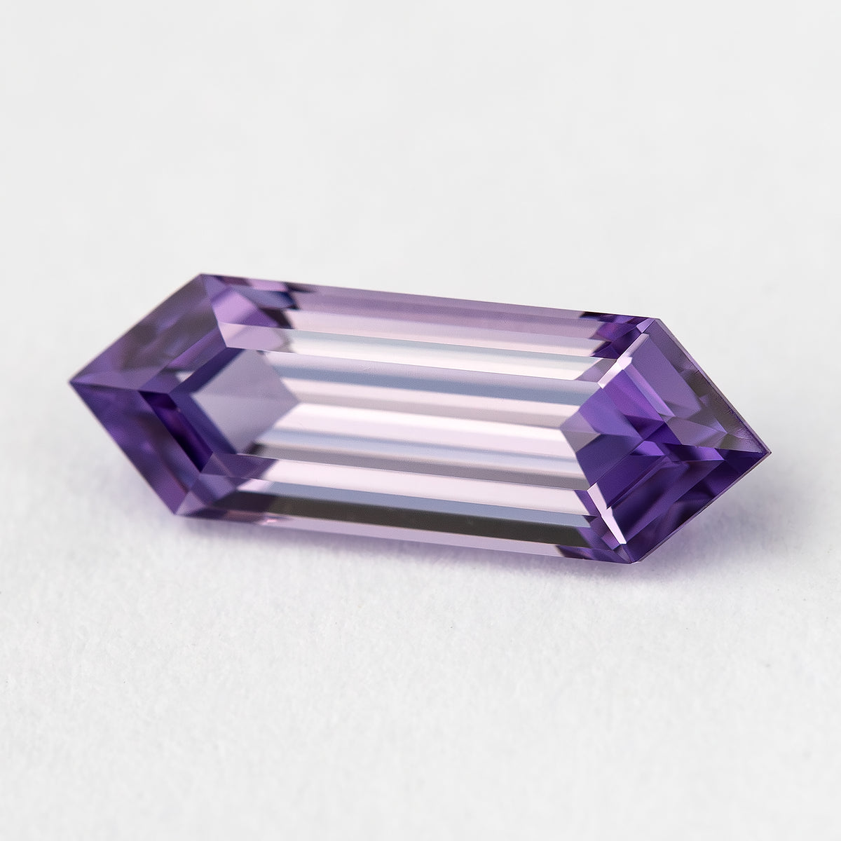 Angled lavender lab-grown sapphire on a pale surface, showing long pale facets and violet pointed ends.