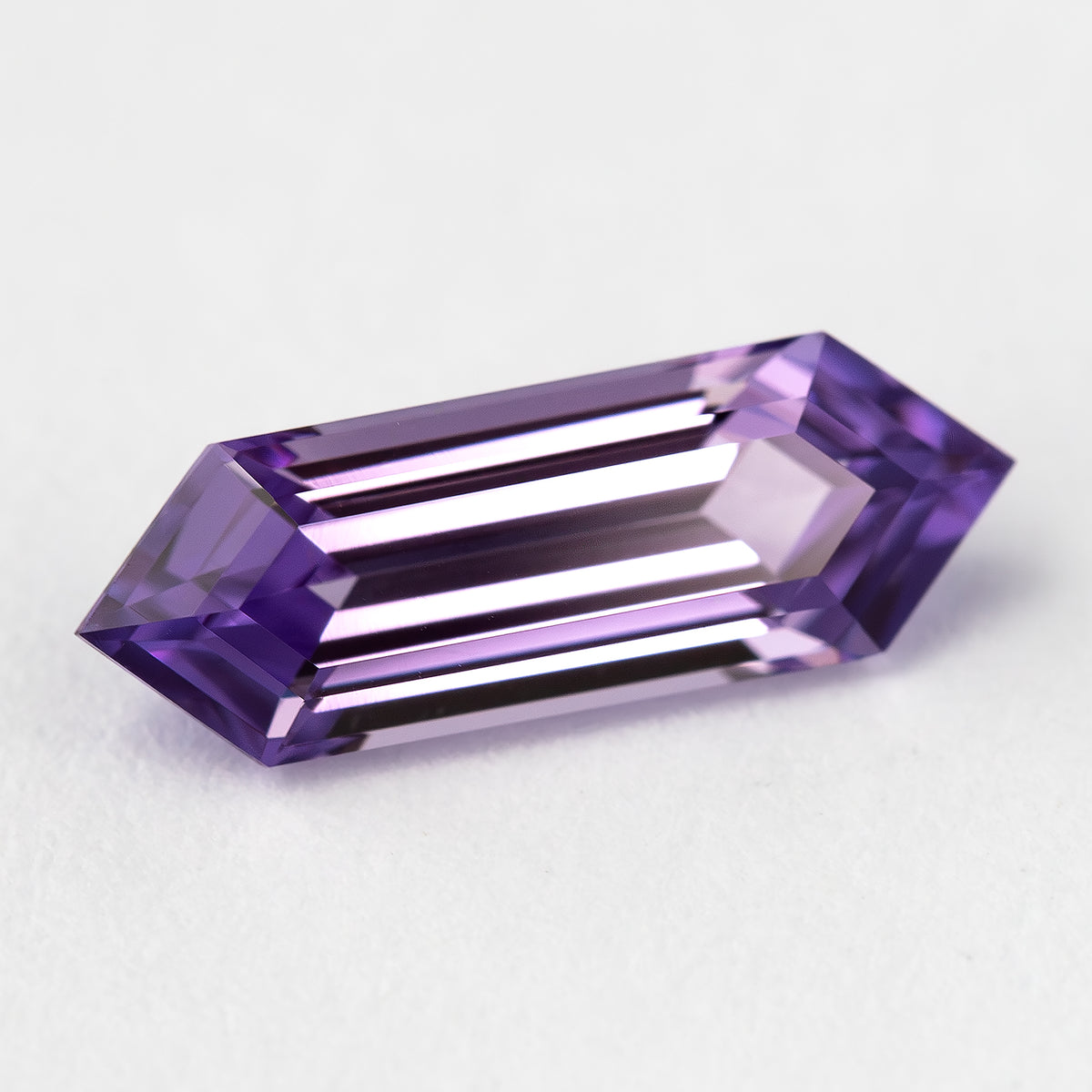 Lavender lab-grown sapphire on a pale surface, showing bright parallel reflections across its long facets.