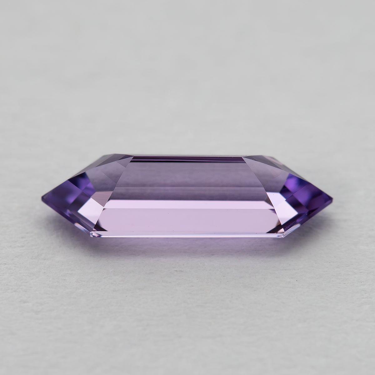 Horizontal view of a lavender lab-grown sapphire on a pale surface, showing a broad pale centre and pointed ends.