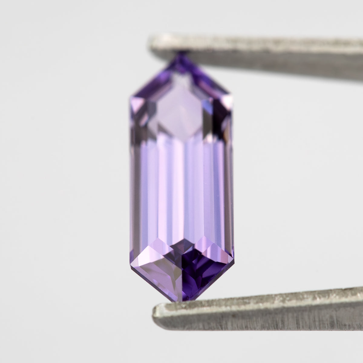 Lavender lab-grown sapphire held vertically between tweezers, with a pale centre and violet pointed ends.