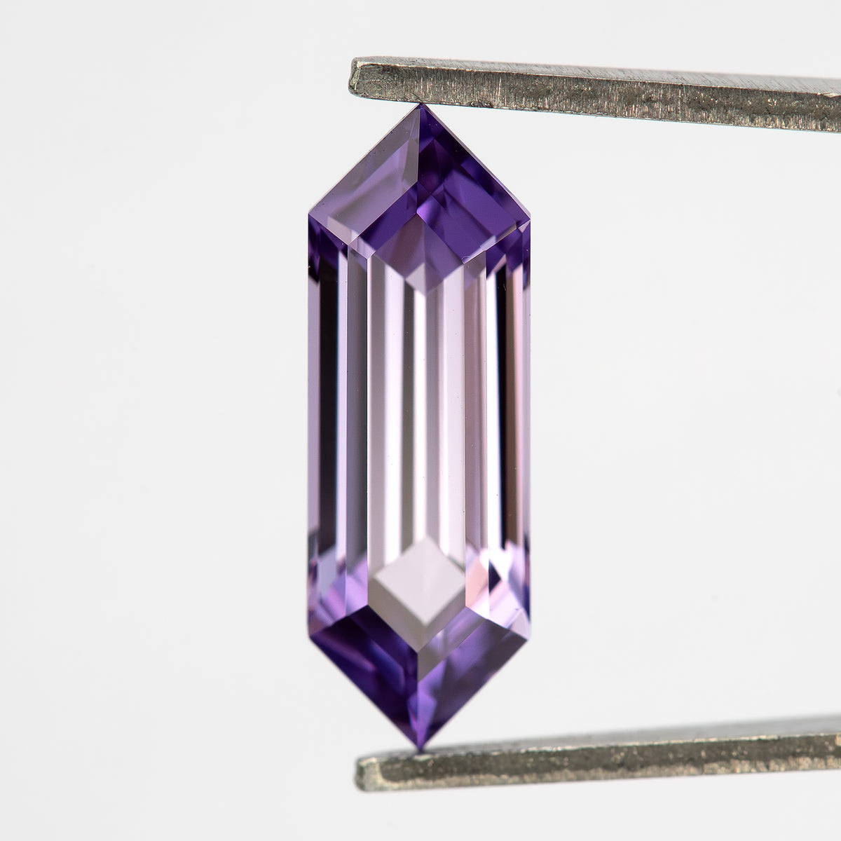 Lavender pink-violet lab-grown sapphire held upright between tweezers, showing its elongated hexagonal shape.