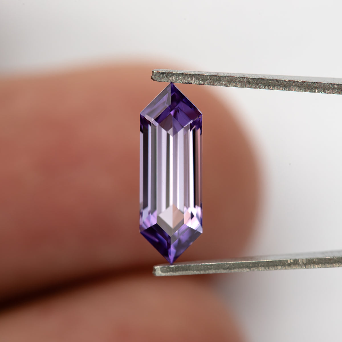 Lavender lab-grown sapphire held upright between tweezers against a fingertip for scale.