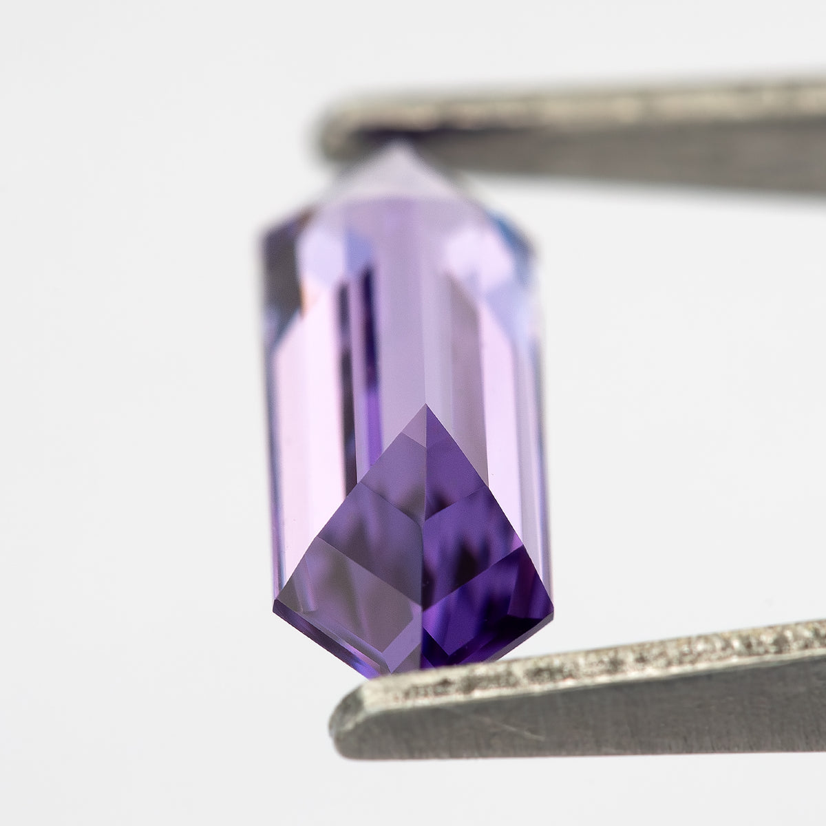 Angled lavender lab-grown sapphire between tweezers, showing pale facets and deep violet reflections near one end.