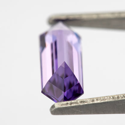 Angled lavender lab-grown sapphire between tweezers, showing pale facets and deep violet reflections near one end.