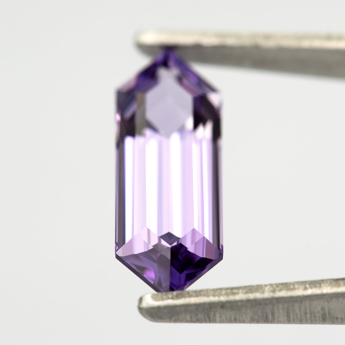 Lavender lab-grown sapphire between tweezers, showing bright central reflections and violet edge facets.