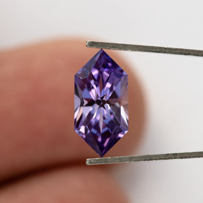 Face-up violet lab-grown sapphire in tweezers against fingers, providing a sense of its size.