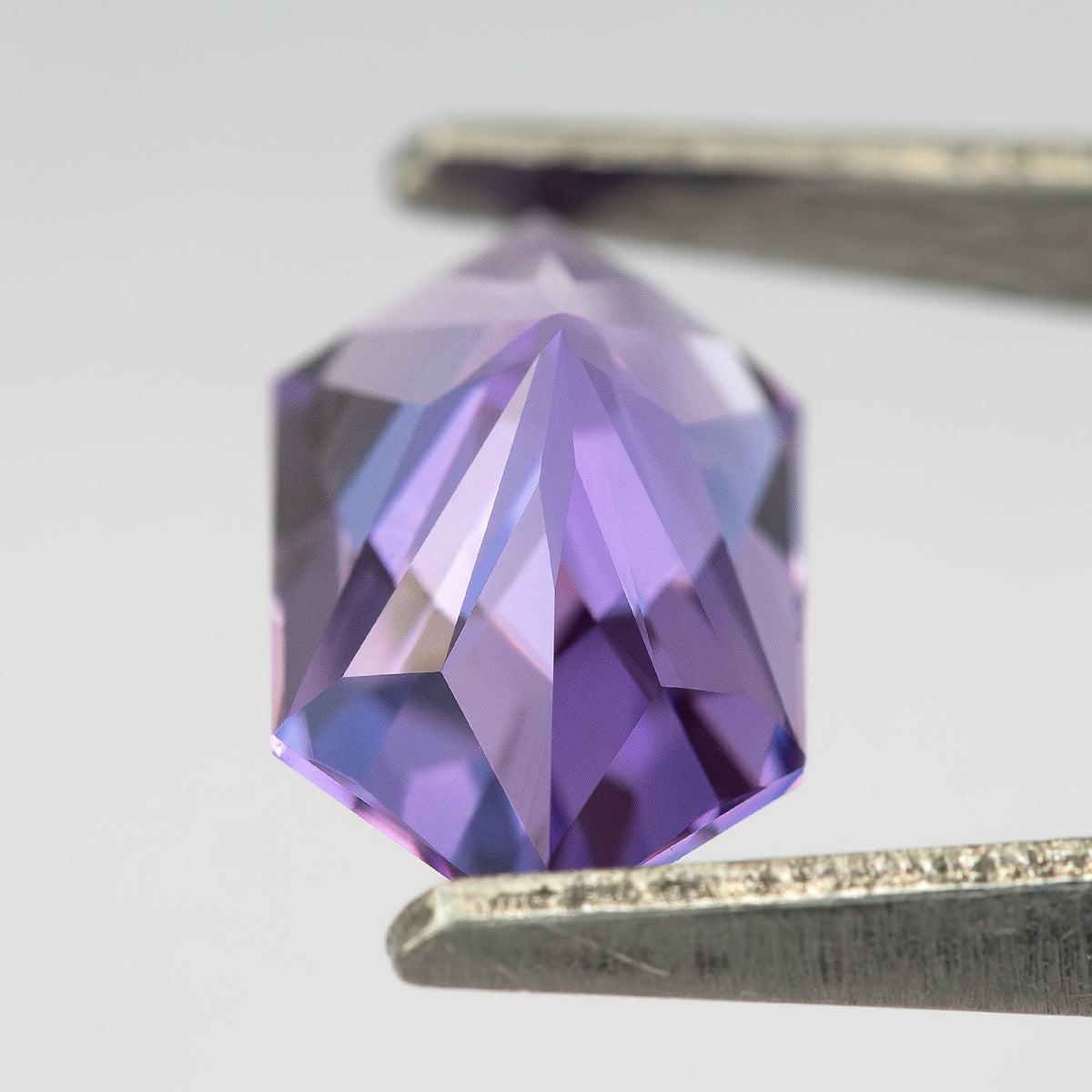 Violet elongated hexagon sapphire held in tweezers with reverse facets visible against fingers.