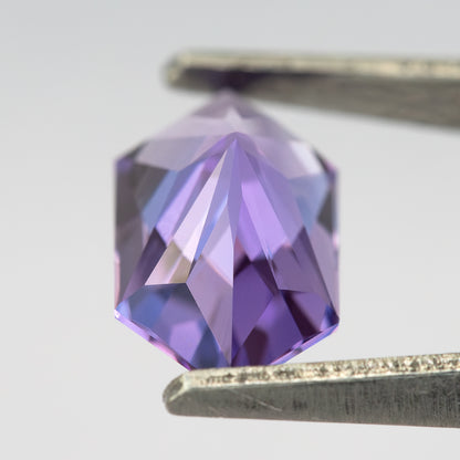 Violet elongated hexagon sapphire held in tweezers with reverse facets visible against fingers.