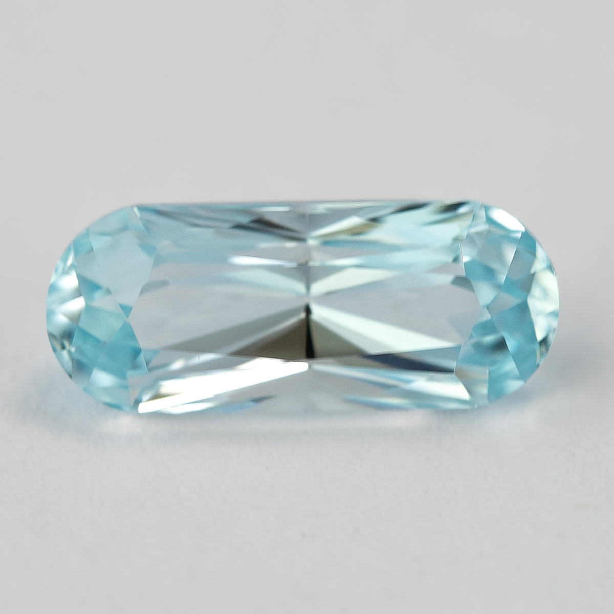 Light blue long-oval sapphire on a pale surface, with rounded ends and elongated facet reflections.