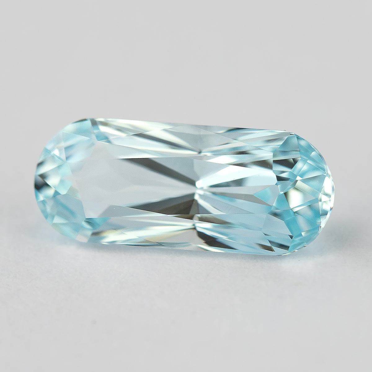 Light blue long-oval sapphire tilted on a pale surface, showing the edge and crown.