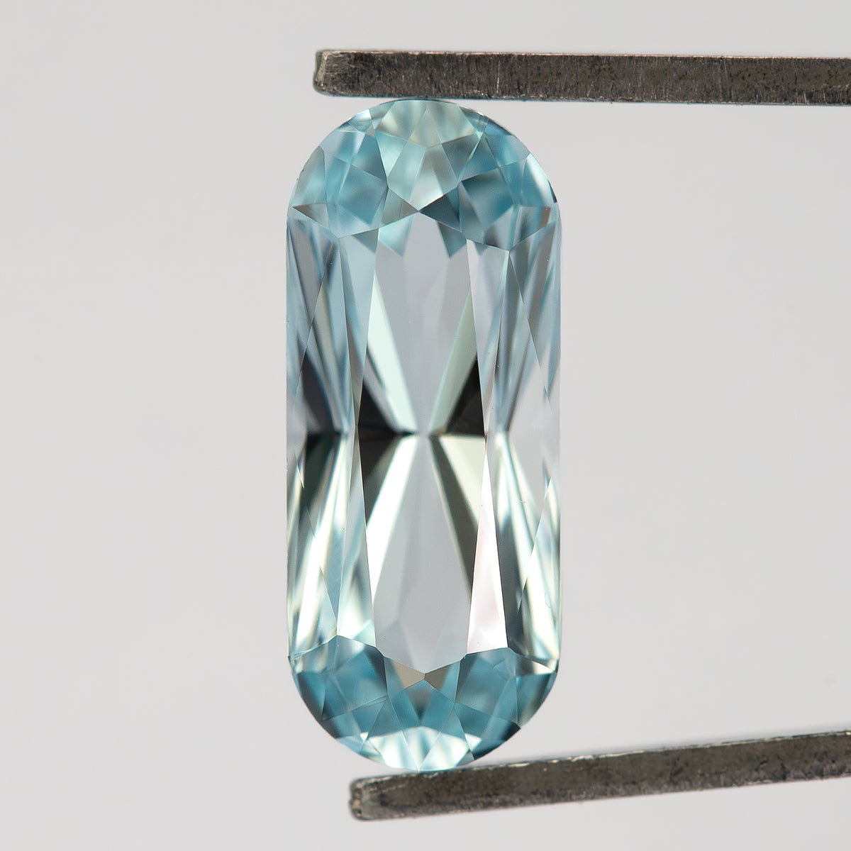 Light blue long-oval sapphire held crown-side toward the camera in metal tweezers, with an elongated table and rounded ends.