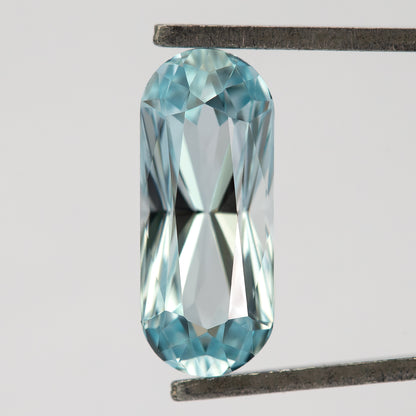 Light blue long-oval sapphire held crown-side toward the camera in metal tweezers, with an elongated table and rounded ends.